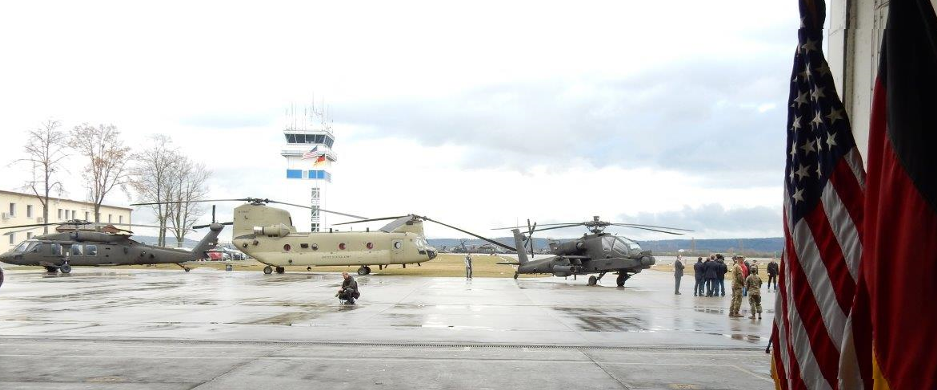 Zwei Kampfhubschrauber AH-64E-Apache und ein Transporthubschrauber CH-47F Chinook auf dem Flugfeld vor dem Kontrollturm.